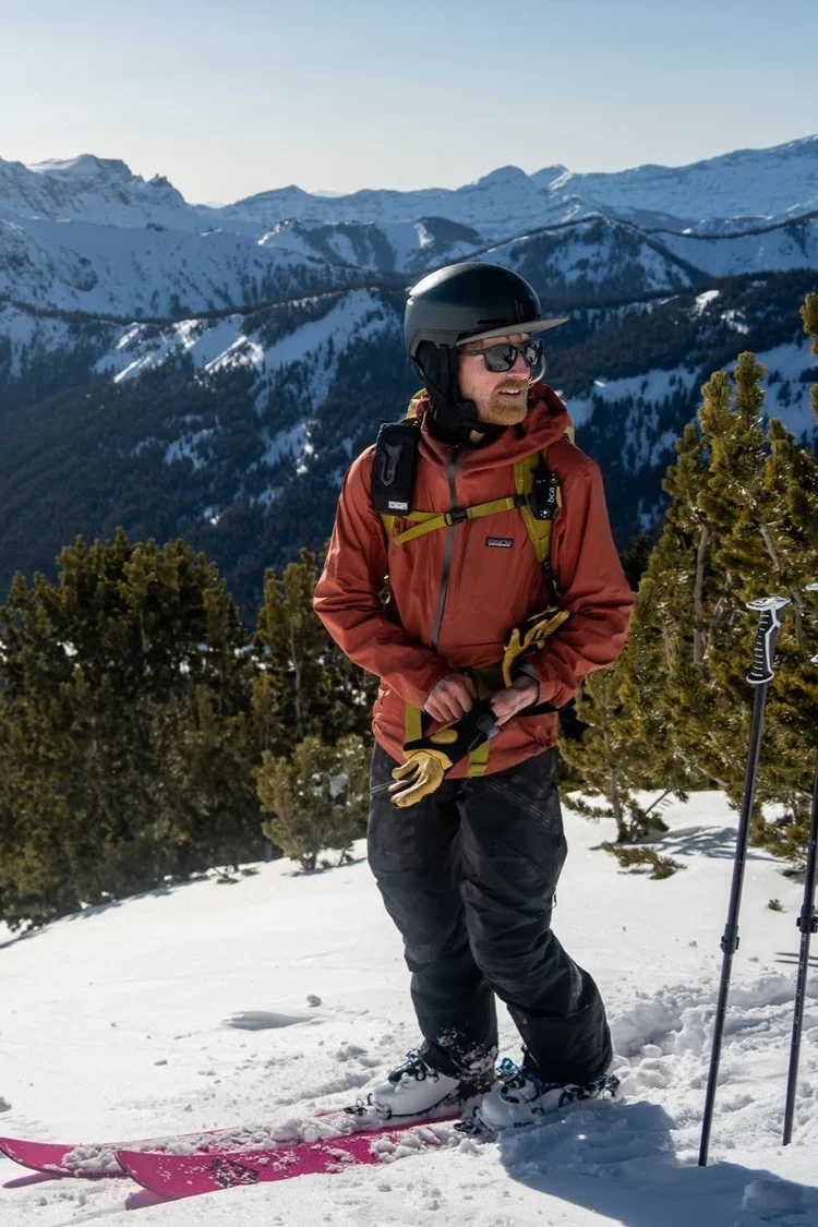 Stephen Gill in winter gear standing in snowy mountain terrain, holding a ski pole with skis resting on the snow beside him