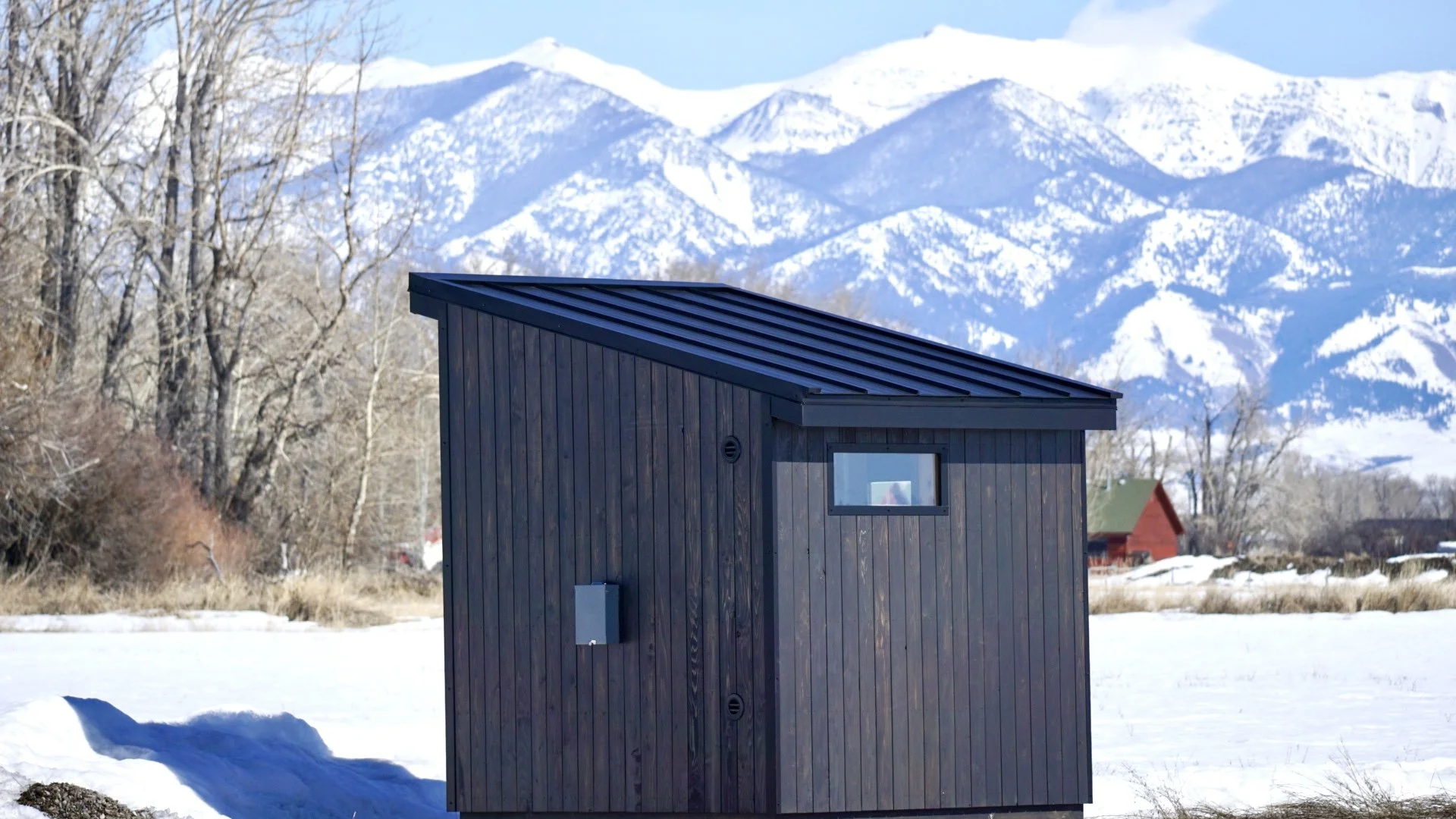 Blackened-cedar sauna with a standing-seam roof set against snow-covered Montana mountains