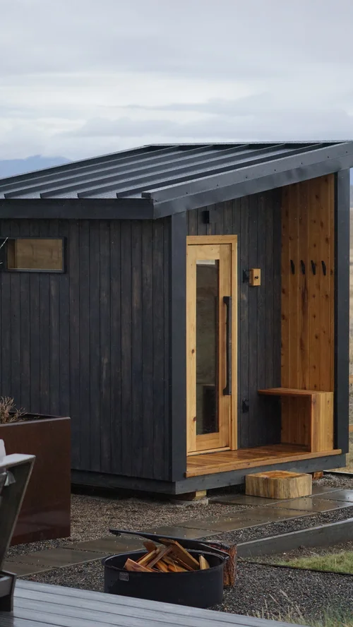 Black sauna with a warm cedar entry porch, bench, and fire pit on a stone patio