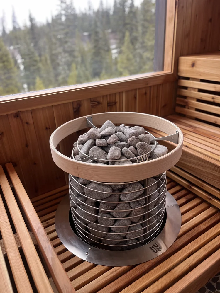 Huum heater stacked with rounded river stones beside tiered cedar benches and a forest window