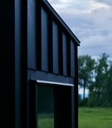 Close-up angled view of the black corrugated-metal sauna exterior, showing its sloped roofline and a window against green grass and trees at dusk